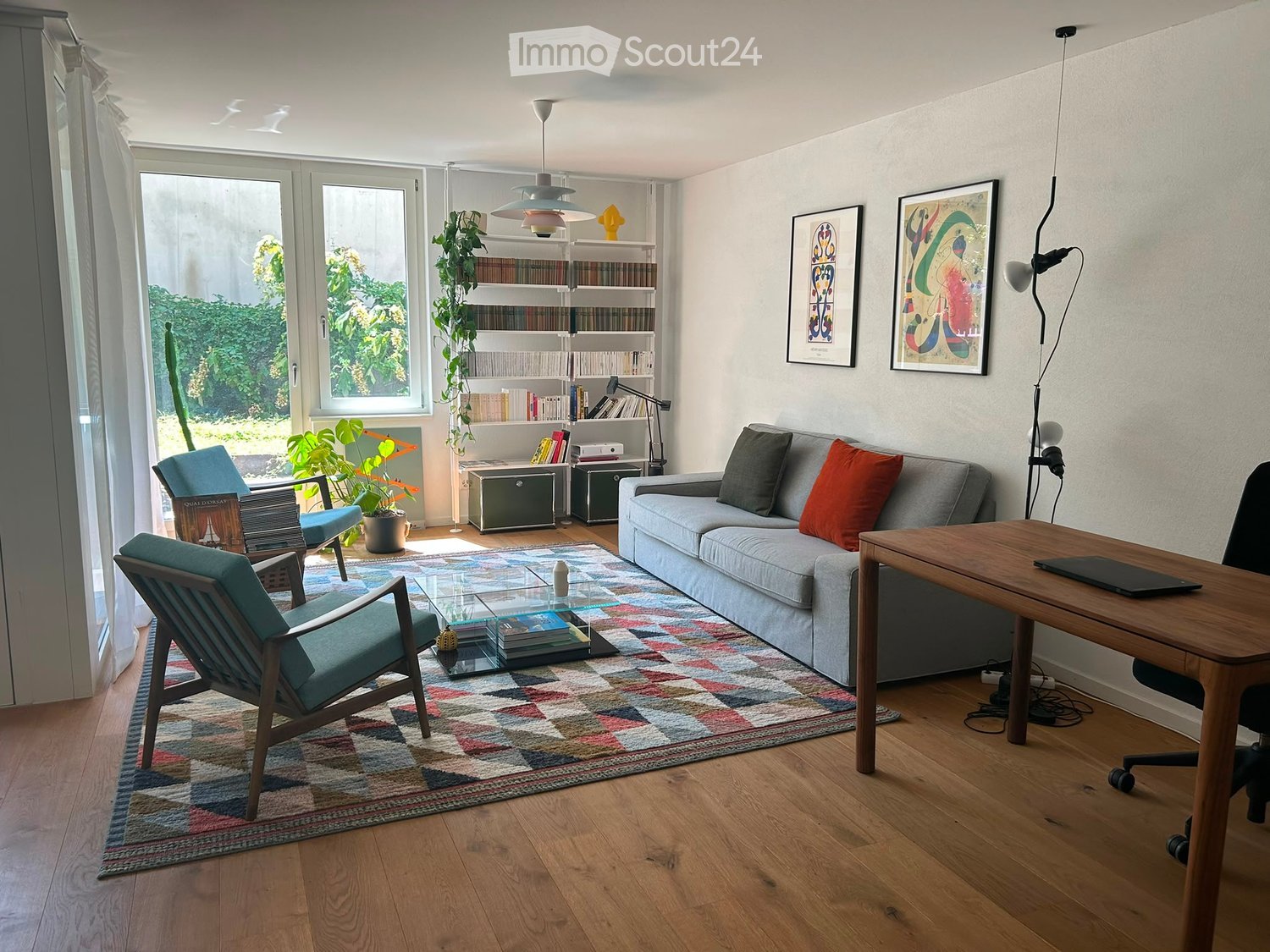 Modern living room with a grey sofa, wooden chairs, a glass coffee table, a wooden desk with a laptop, a bookshelf, and decorative plants.