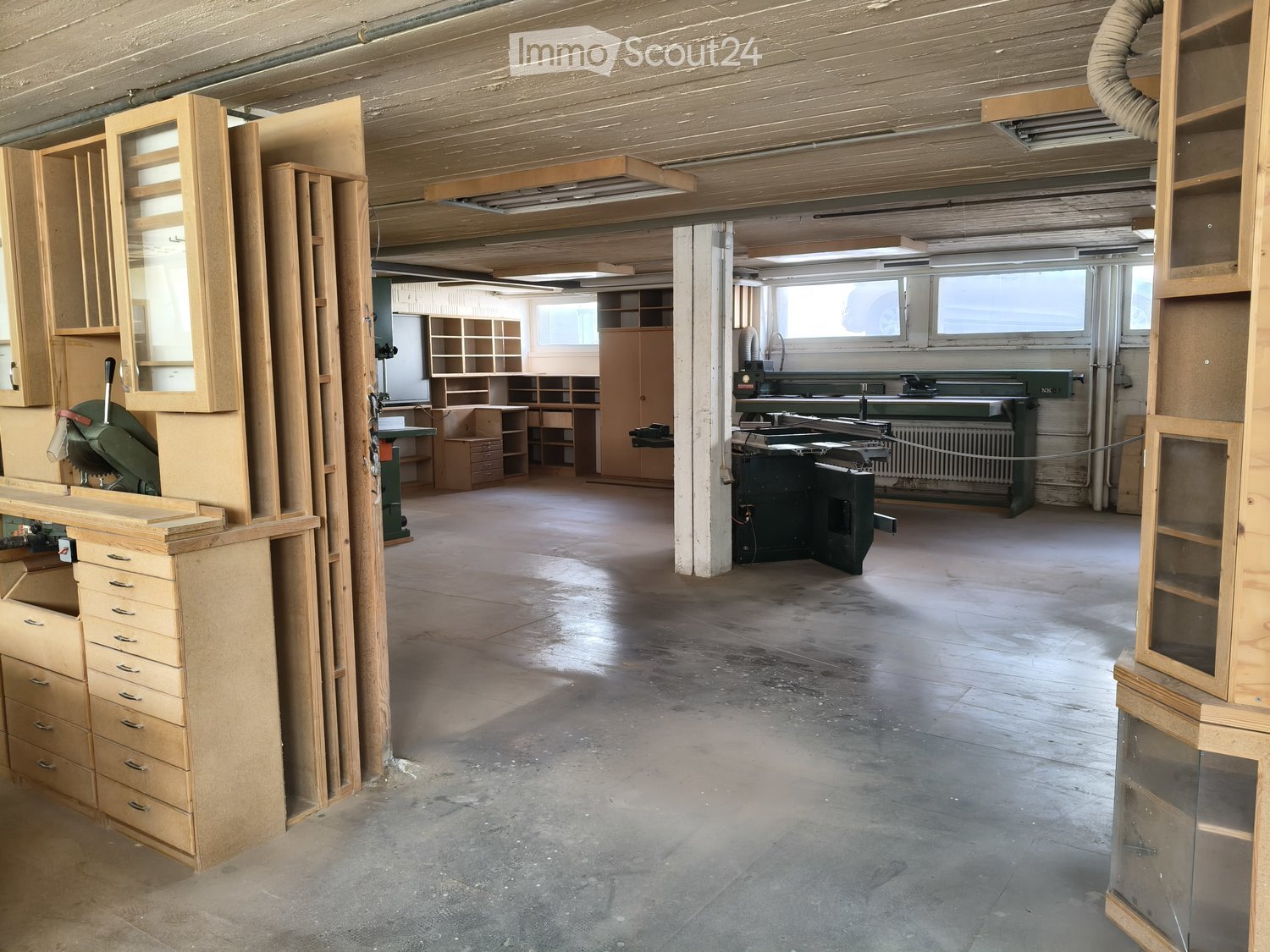 Empty workshop with exposed wooden beams, concrete floor, and various wooden furniture pieces and tools stacked around the space.