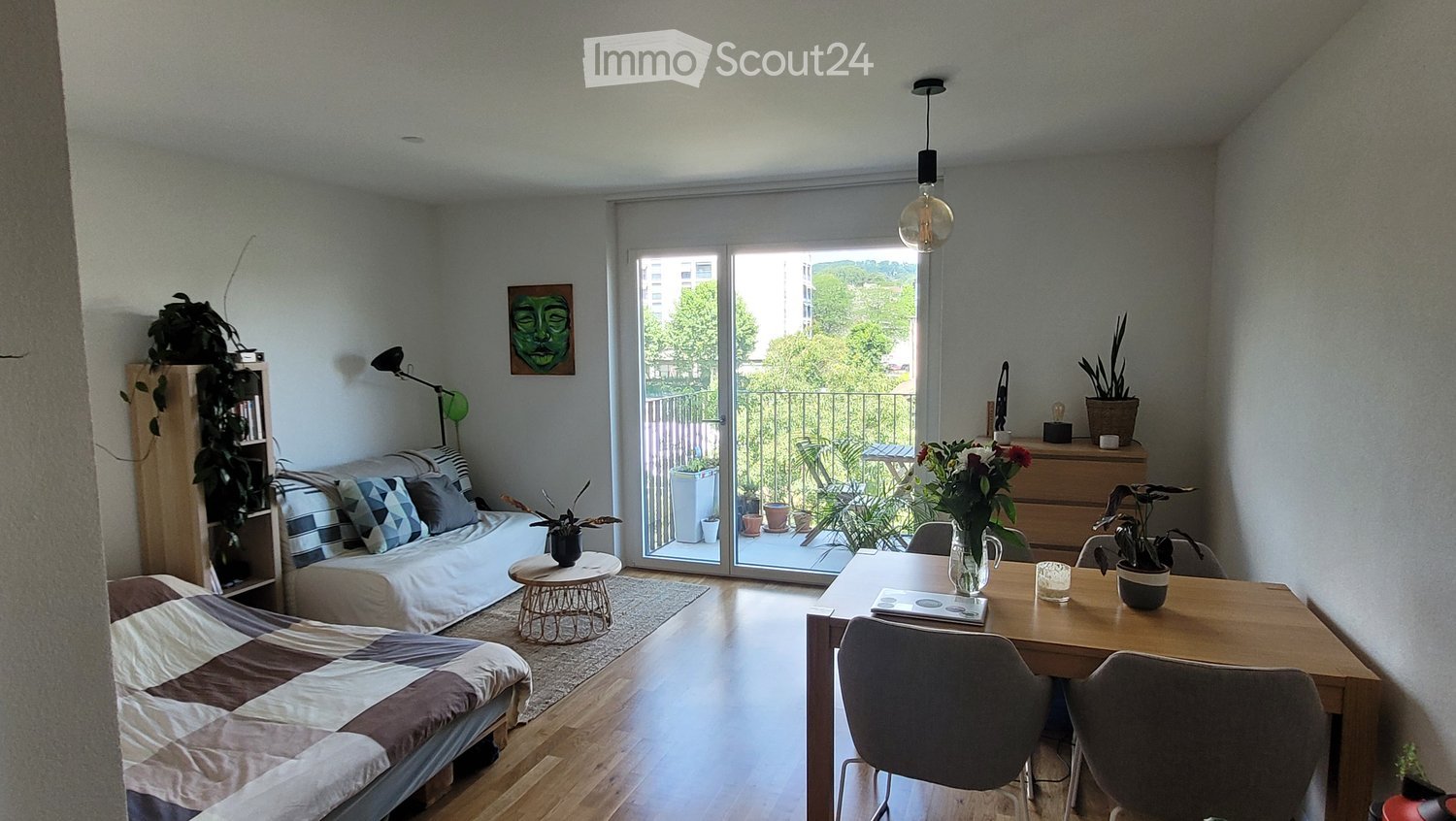 A modern, well-lit living space with a sofa, bed, dining table, chairs, bookshelf, and various decorative plants. Large windows and a sliding glass door lead to a balcony.