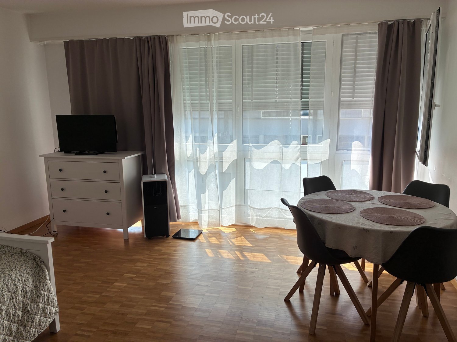 A modern, well-lit room with wooden flooring, large windows with blinds and curtains, a dining table with chairs, a TV on a stand, and an air conditioner unit.