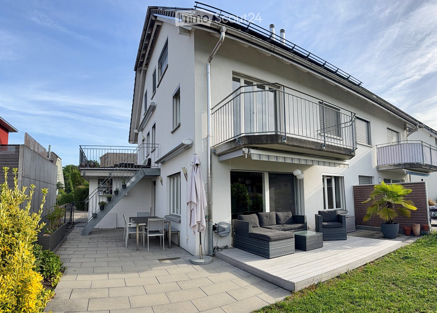 Modern outdoor patio area with a sofa, ottoman, chairs, a table, and an umbrella. The patio is adjacent to a two-story house with balconies and a staircase leading to an upper terrace.