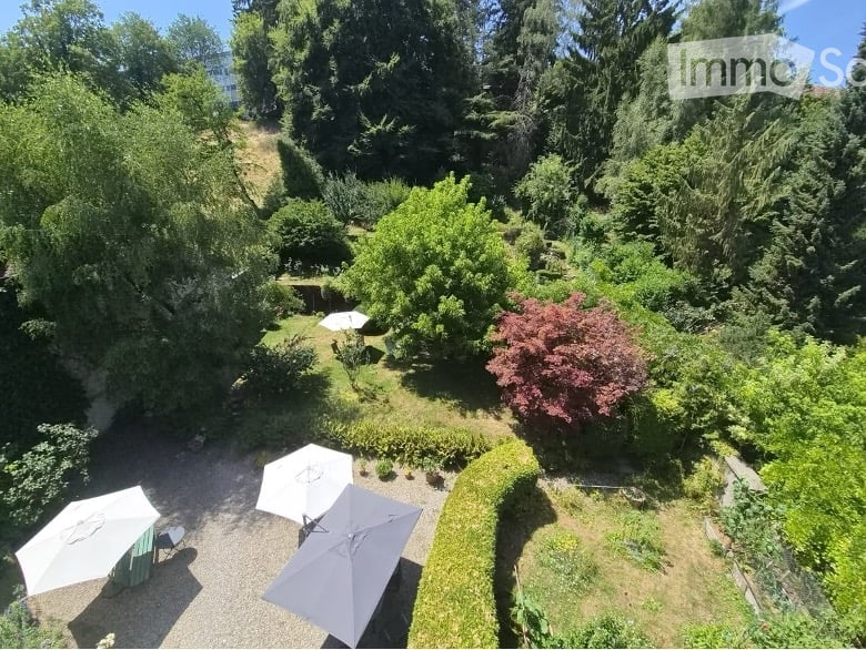 A lush garden area with various trees, shrubs, and a paved patio with several white umbrellas.