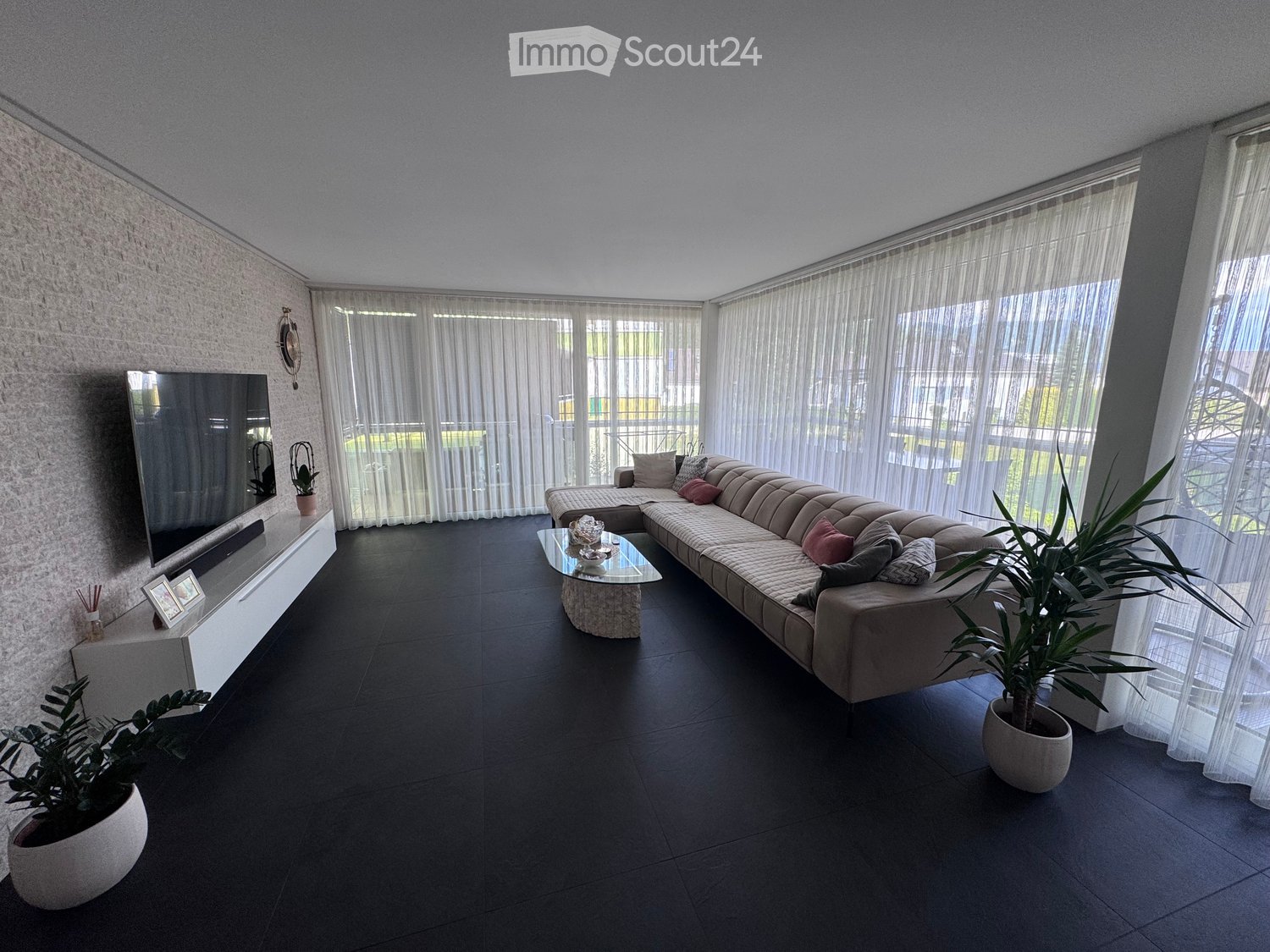 Modern living room with a large sectional sofa, glass coffee table, flat-screen TV on a white console, and potted plants.