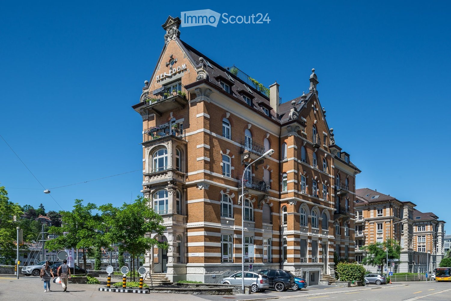 A large, ornate brick building with multiple stories, arched windows, decorative facades, and a prominent roofline. The building features balconies and is situated on a street with parked cars and pedestrians.