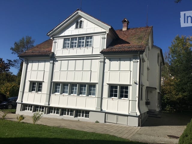 A two-story white house with a red-tiled roof, multiple windows, and a small chimney. The house is surrounded by a well-maintained lawn and trees.
