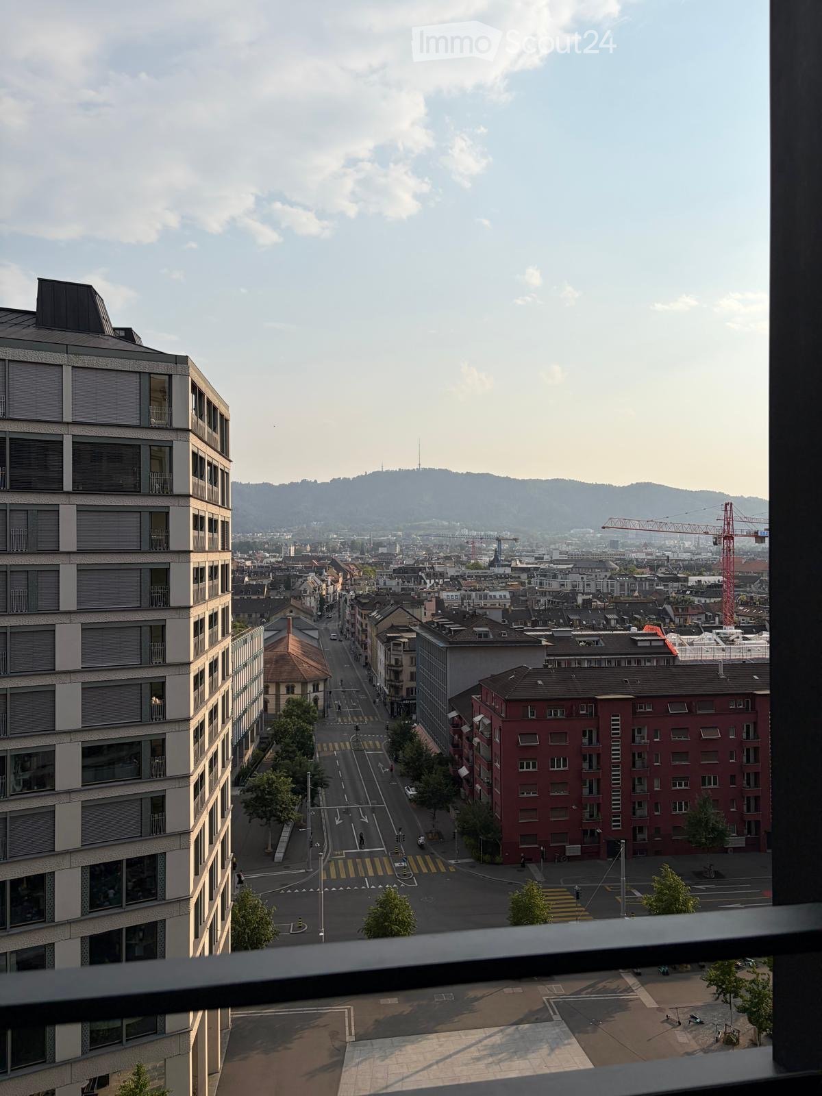 Balcony view overlooking a city street with buildings, traffic, and distant hills.
