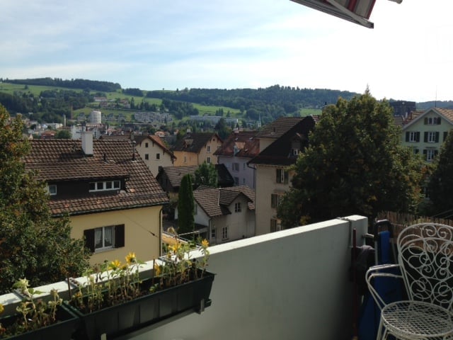 Balcony with a scenic view of a town and surrounding hills, featuring potted plants and a white metal chair.