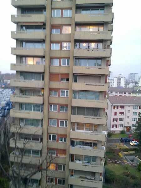 A tall, multi-story residential building with numerous windows and balconies, surrounded by other buildings and a parking area.