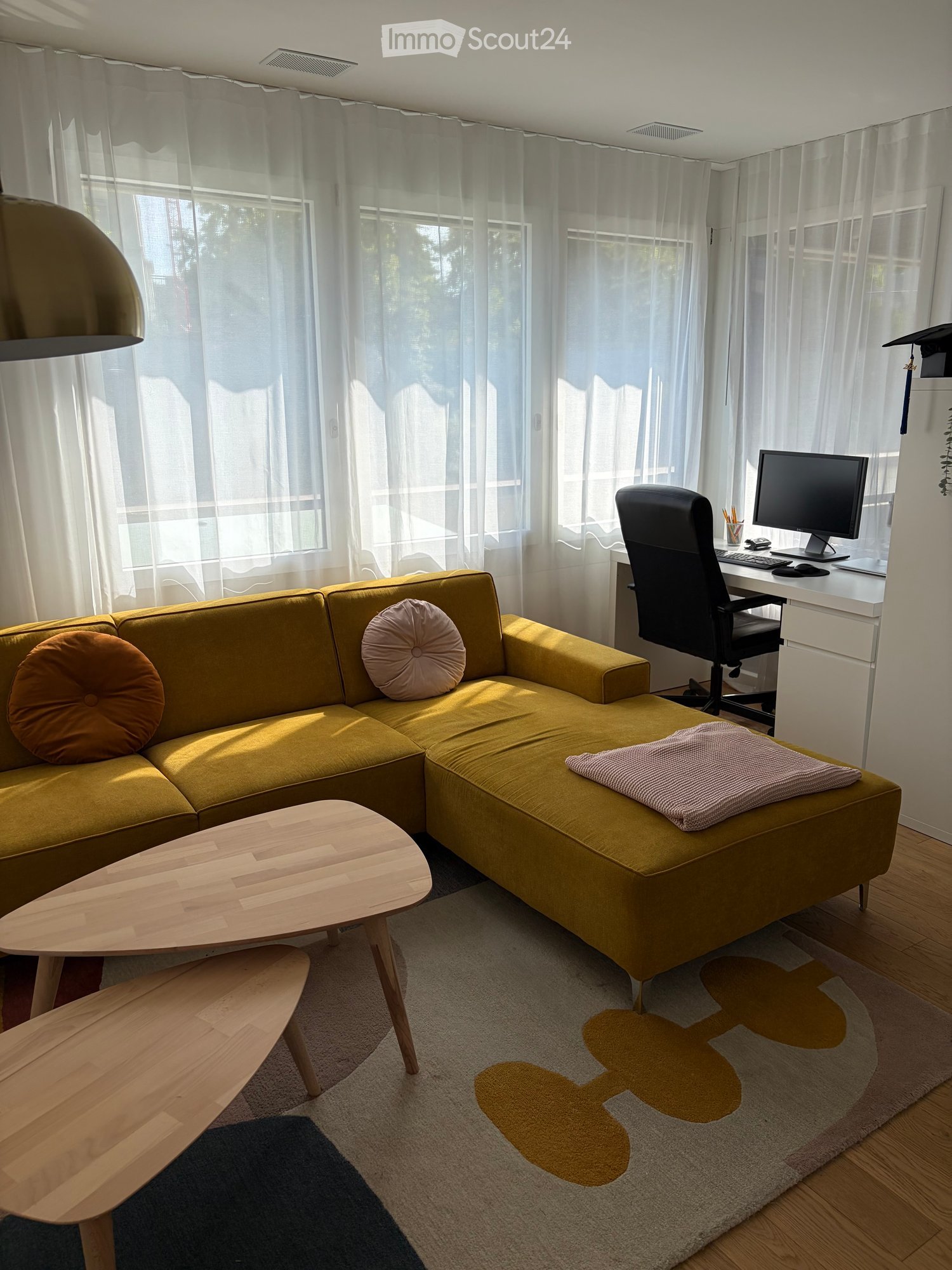 Bright living room with a mustard yellow sectional sofa, wooden coffee table, office desk with computer, and large windows with sheer curtains.