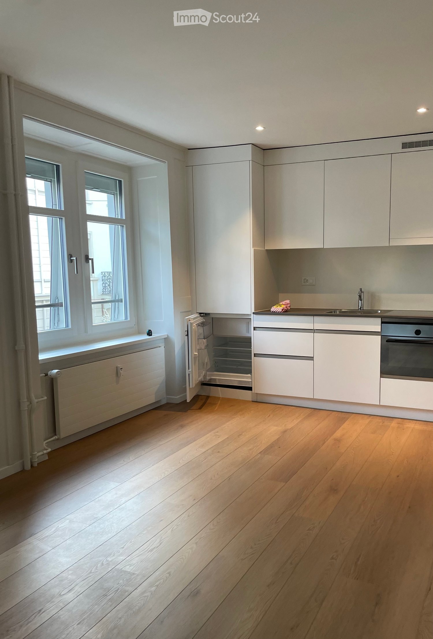 Modern kitchen with light-colored cabinets, wooden flooring, and large windows allowing natural light. Features include a sink, faucet, oven, and refrigerator.
