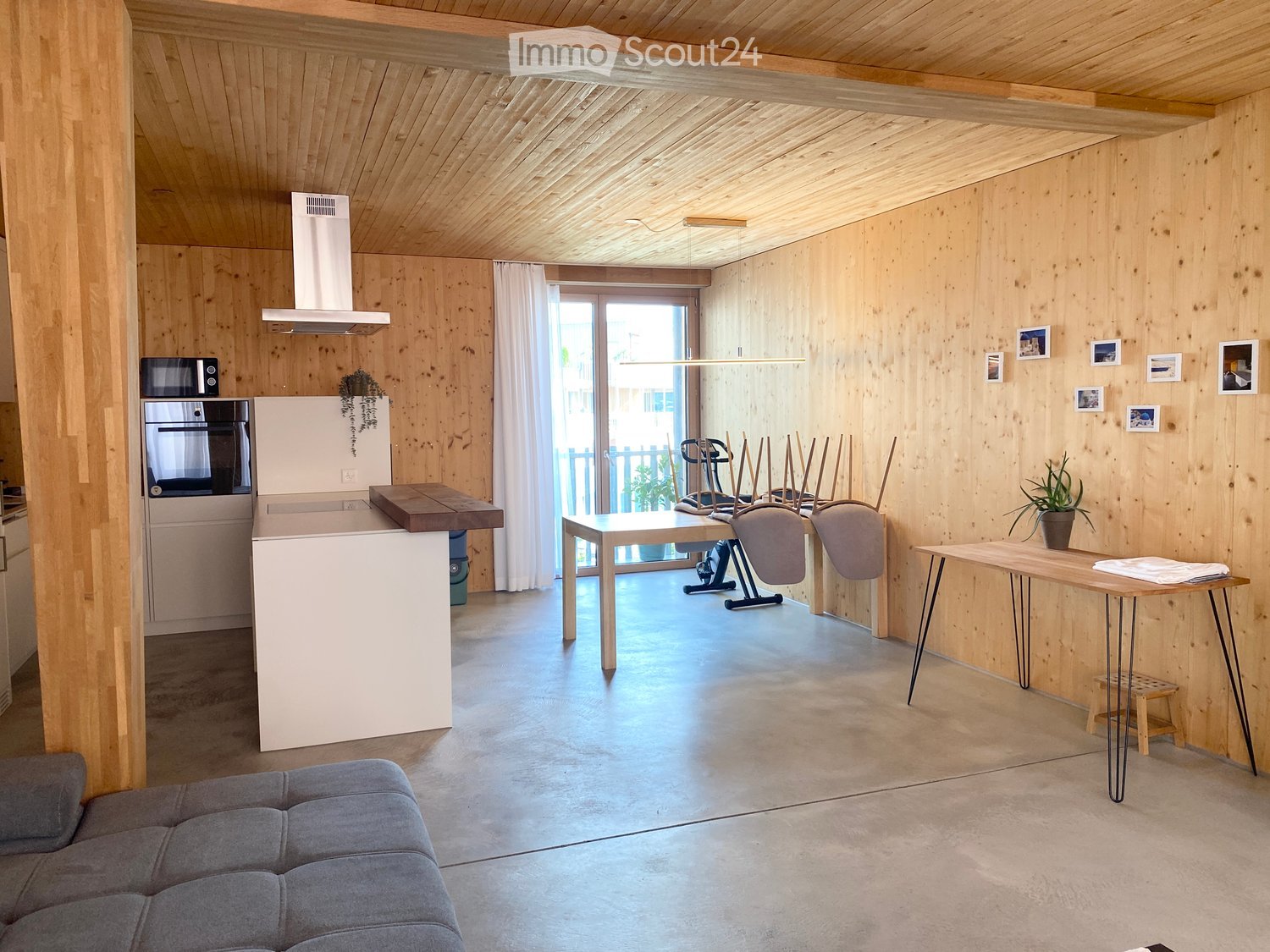 Open-plan living and kitchen area with wooden walls and ceiling, grey sofa, kitchen island, oven, refrigerator, dining table, chairs, and potted plants.