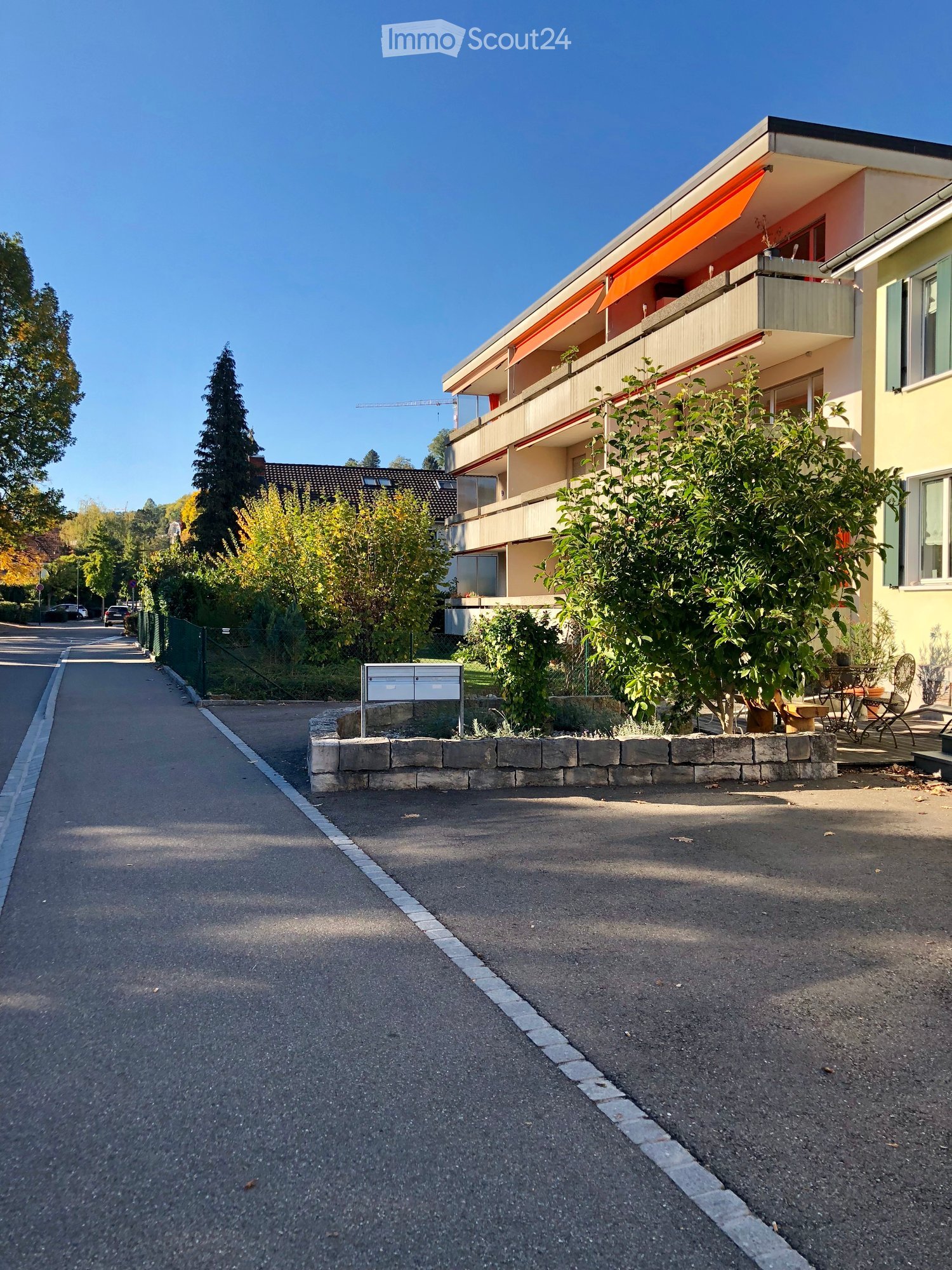 Modern multi-story residential building with balconies, surrounded by greenery and a paved street.