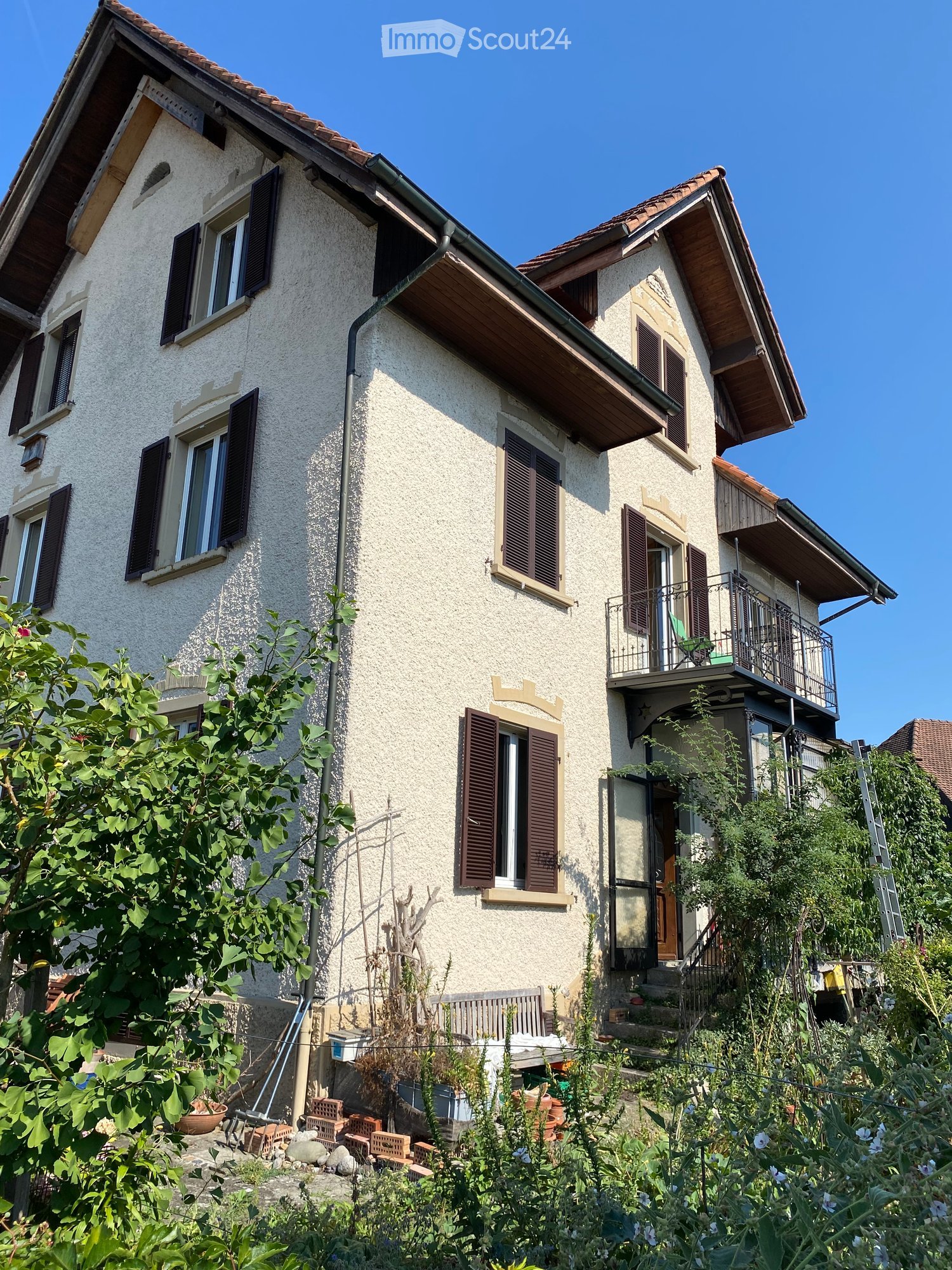 Two-story house with beige stucco exterior, multiple windows with dark shutters, a balcony on the upper floor, and a small garden area with plants and a bench.