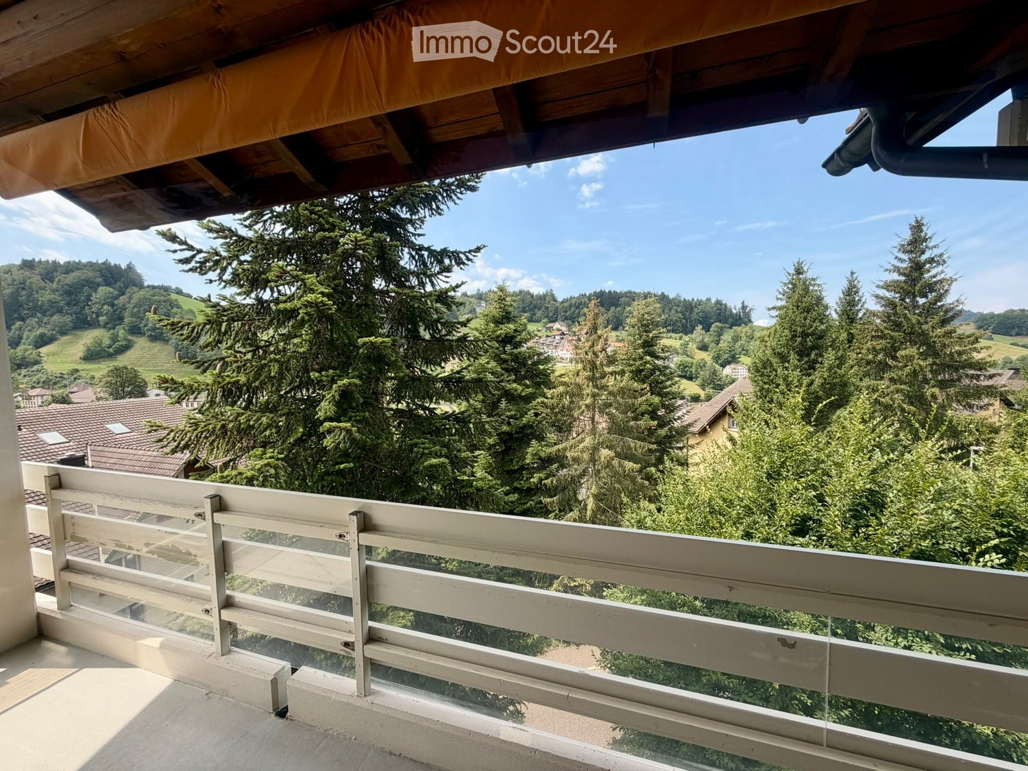 Balcony with wooden railing and glass panels, overlooking a scenic valley with trees and houses.