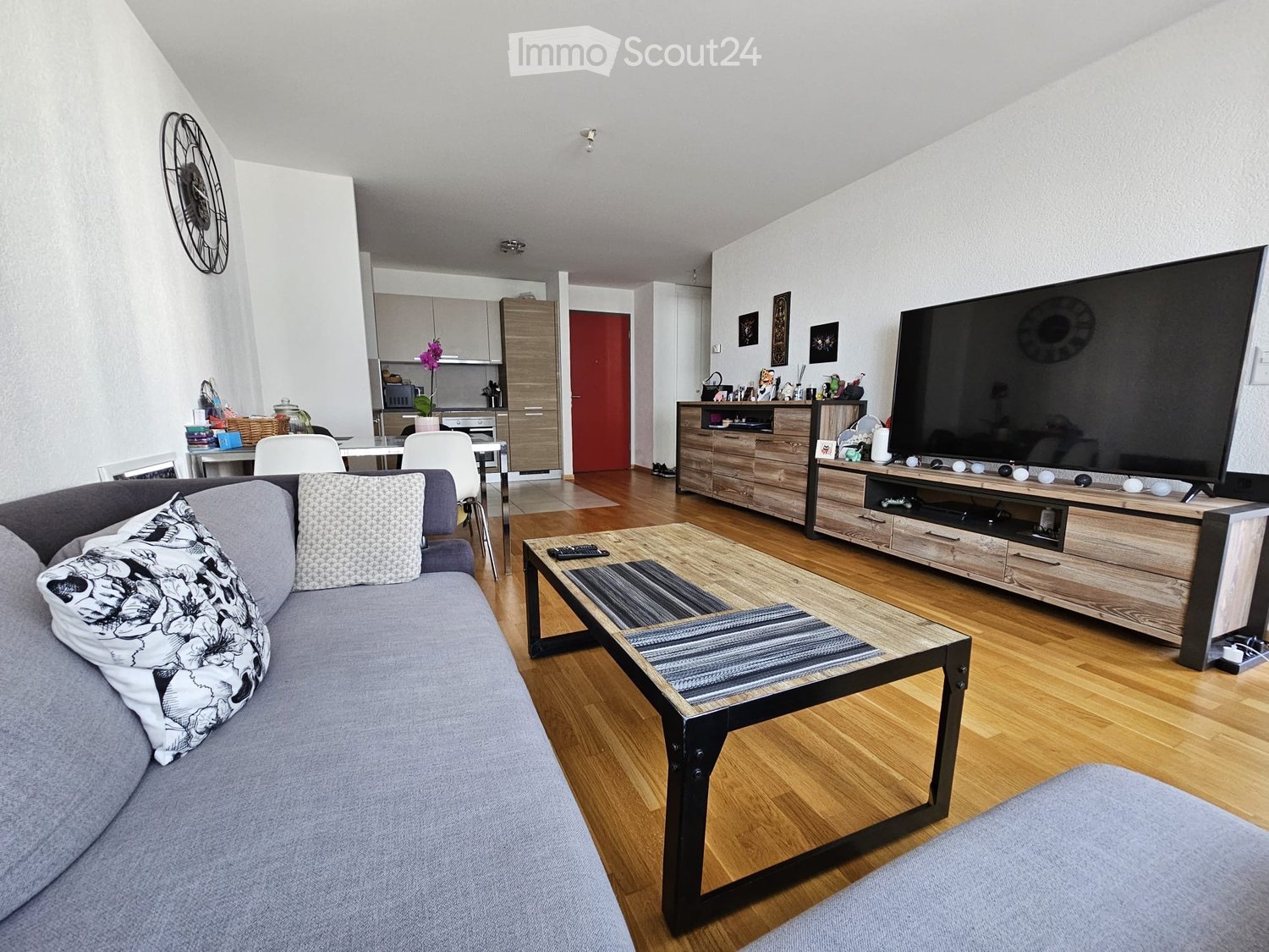 Modern living room with grey sofa, coffee table, TV stand, and dining area with chairs and table. Kitchen area visible with cabinets and appliances.