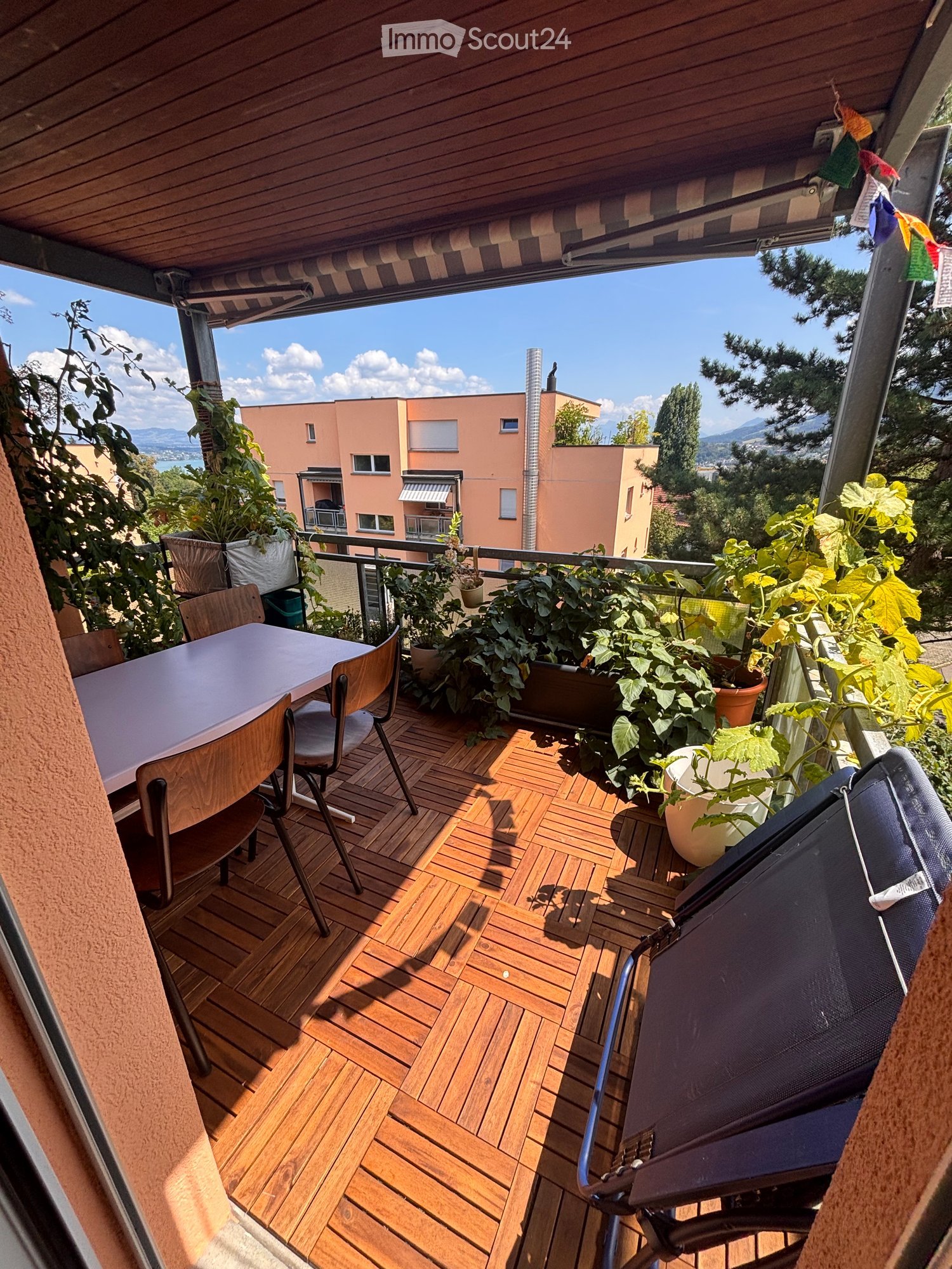 Balcony with wooden decking, table, chairs, and potted plants, overlooking a residential area with buildings and trees.