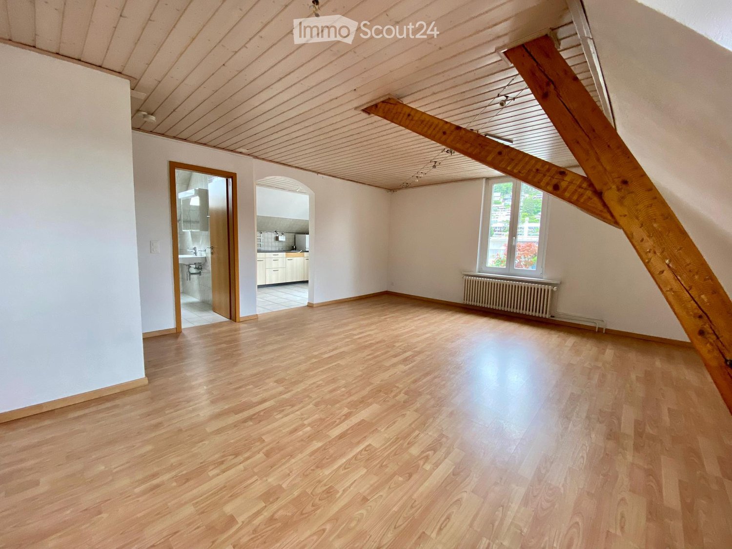 Empty room with wooden beams, wooden floor, windows, doors to the bathroom and kitchen, radiator