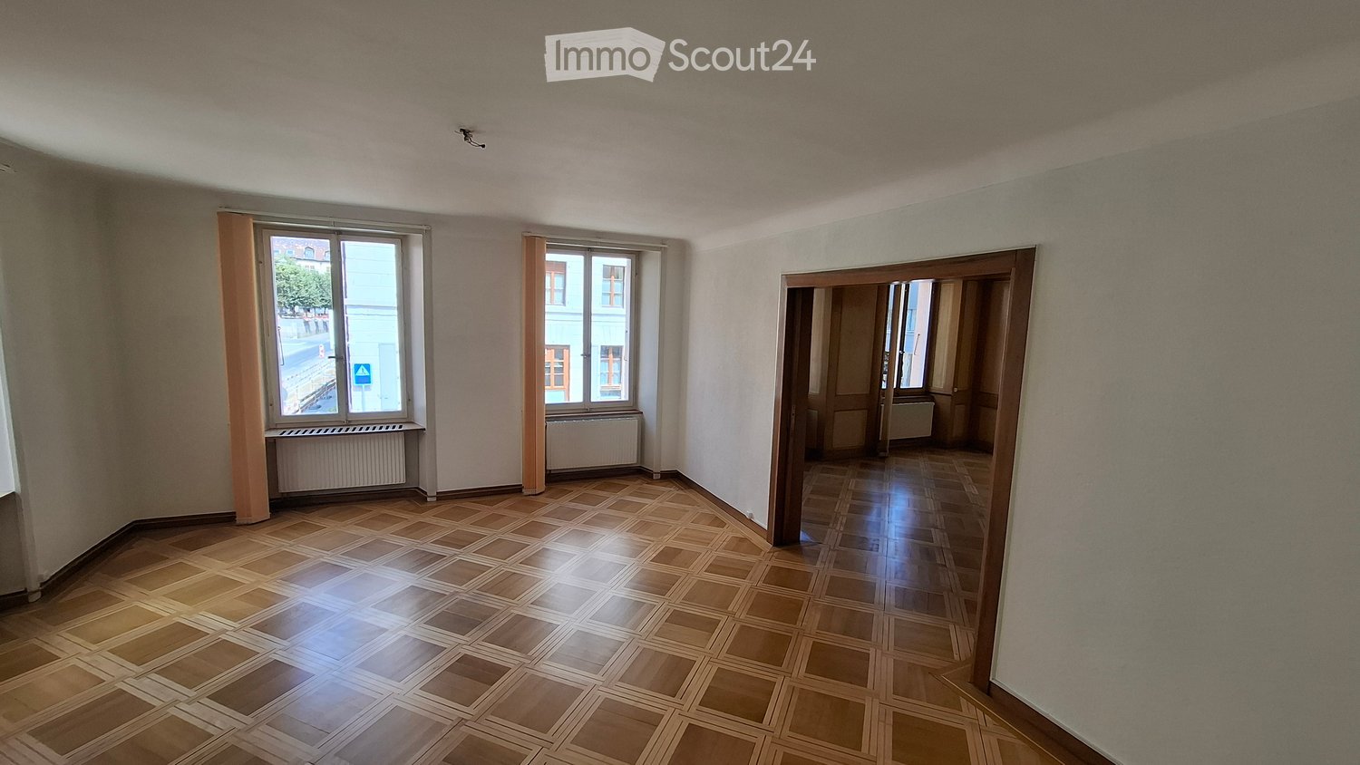 Empty room with wooden flooring, large windows, and a door leading to another room. No furniture or equipment is present.