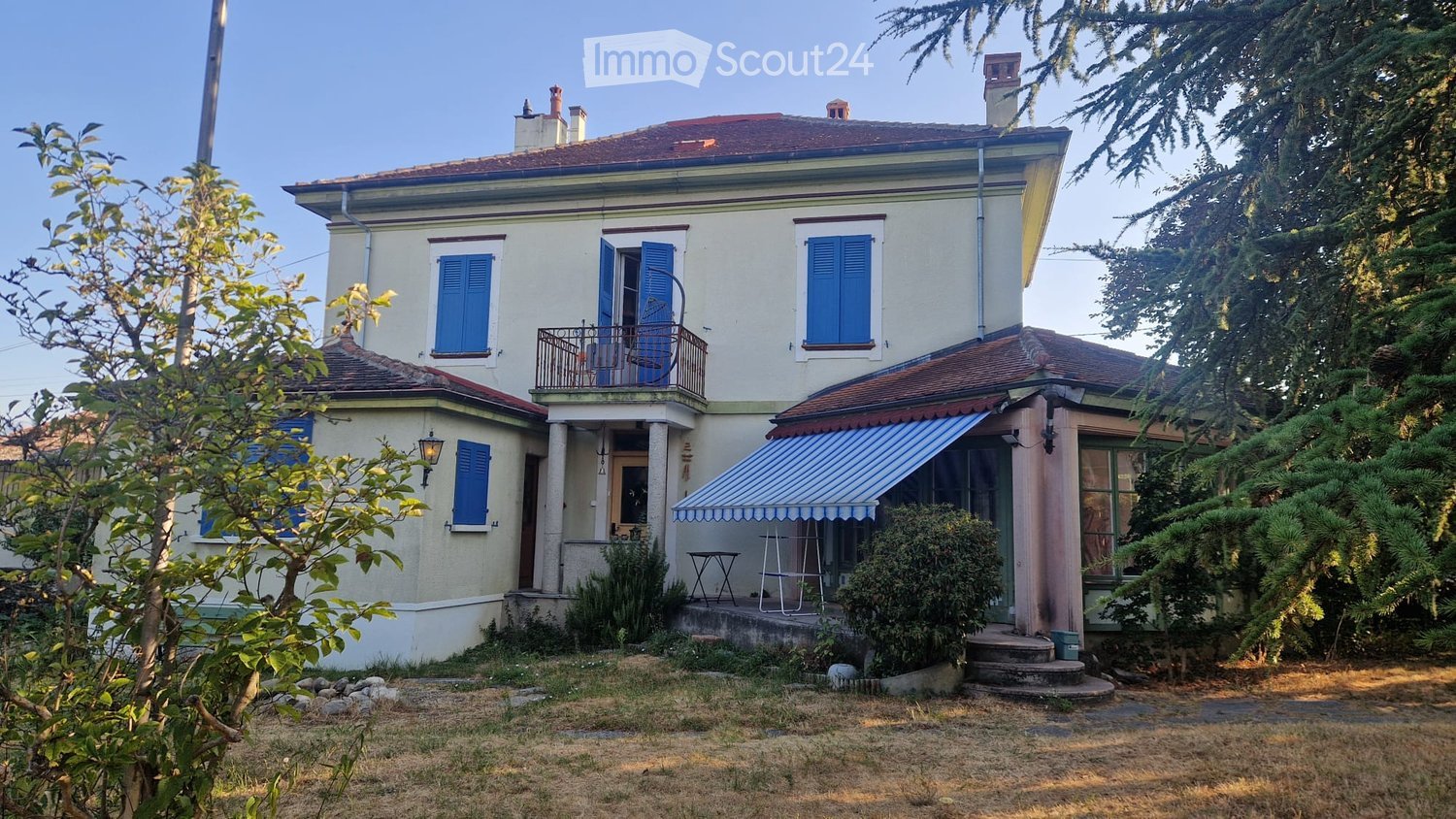 Two-story house with light yellow walls, blue shutters, and a red-tiled roof. Features a balcony with a metal railing and a small awning over the entrance.