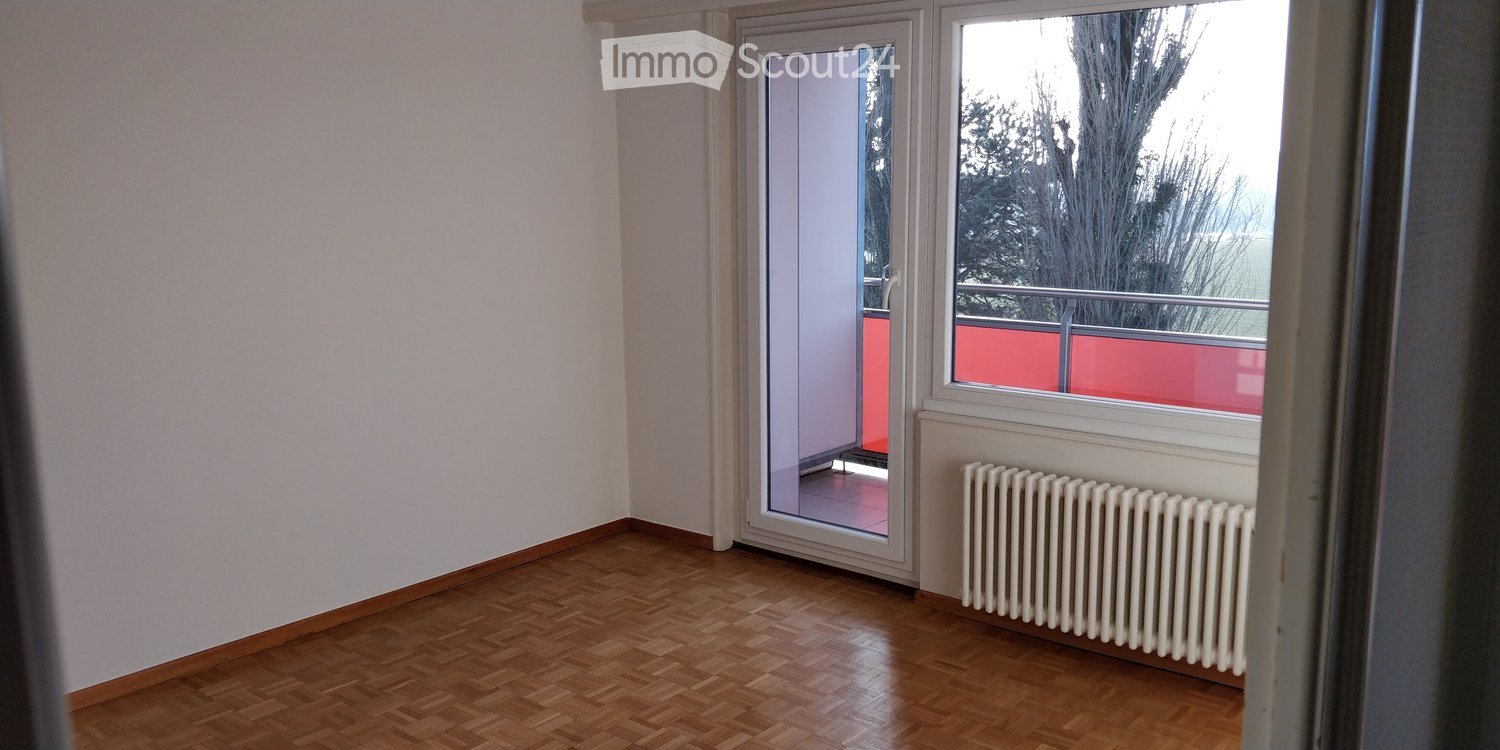 Empty room with wooden flooring, white walls, a radiator, and a sliding glass door leading to a balcony with a red railing.