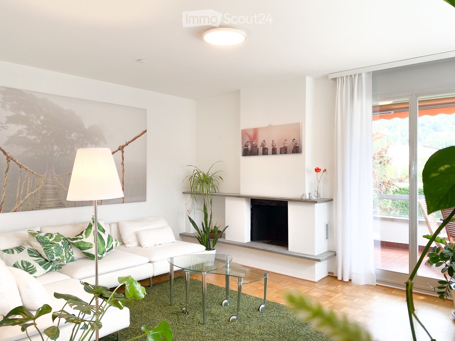 Modern living room with white walls, a white sectional sofa with green cushions, a glass coffee table, a fireplace, and large windows with white curtains.