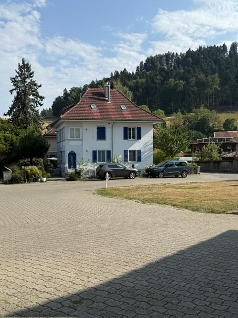 Two-story white house with blue shutters and a red-tiled roof, surrounded by trees and a parking area with two cars.