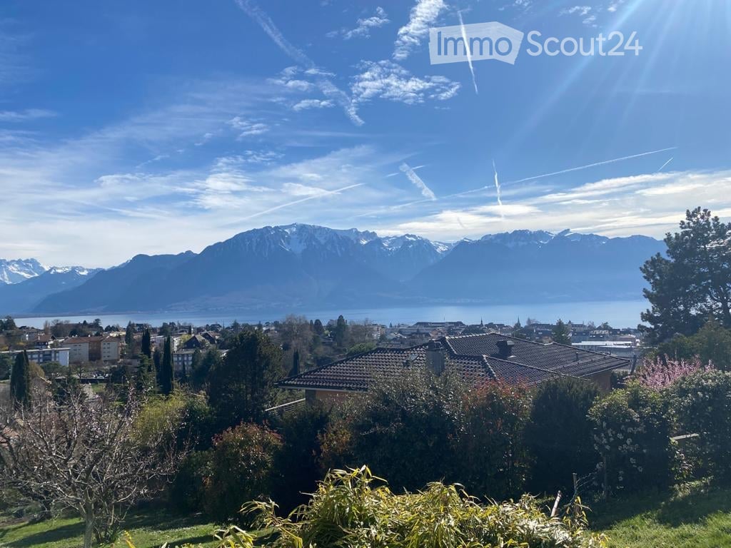 Panoramic view of a cityscape with mountains and a lake in the background, surrounded by trees and greenery.