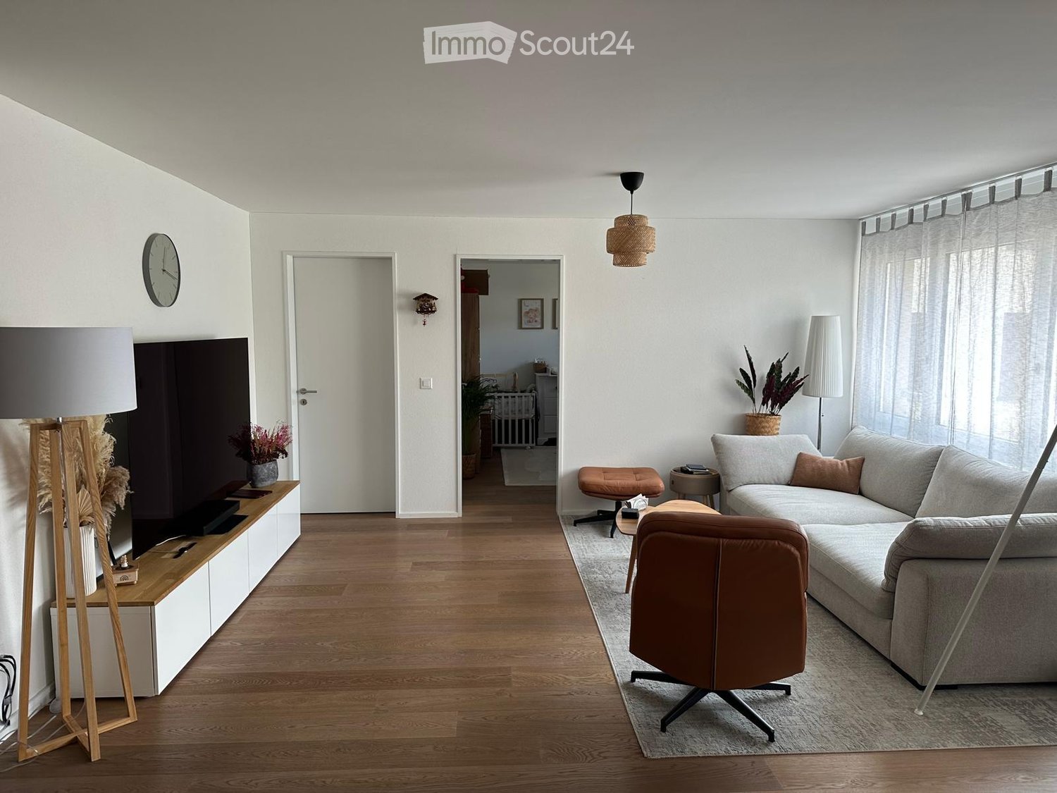Modern living room with light-colored walls, wooden flooring, and large windows. Features a grey sofa, brown leather chair, coffee table, floor lamp, and decorative plants.
