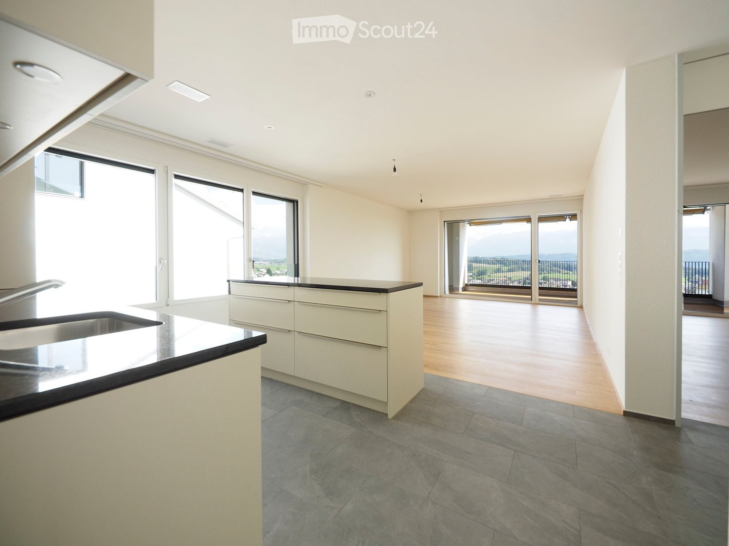 Modern kitchen with white cabinets, black countertop, and stainless steel sink. Large windows and sliding doors lead to a balcony with a scenic view.
