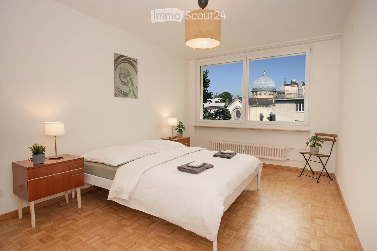 Bedroom with a bed, nightstands, lamps, a chair, a radiator, a window with a view of a dome building, and a picture frame on the wall.