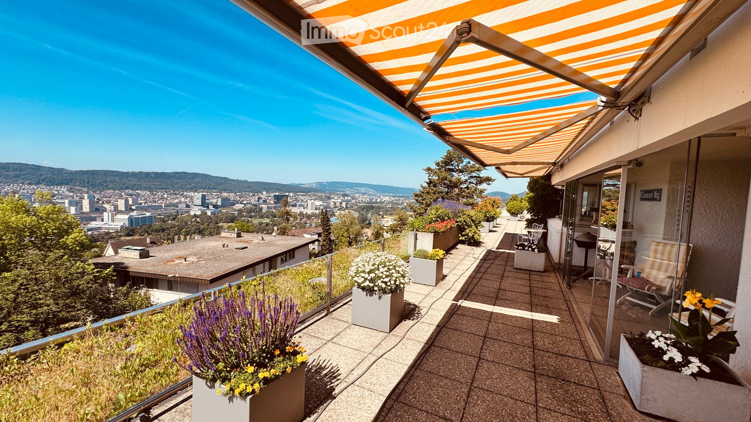 Balcony with a retractable awning, tiled floor, potted plants, and a view of the city and mountains.
