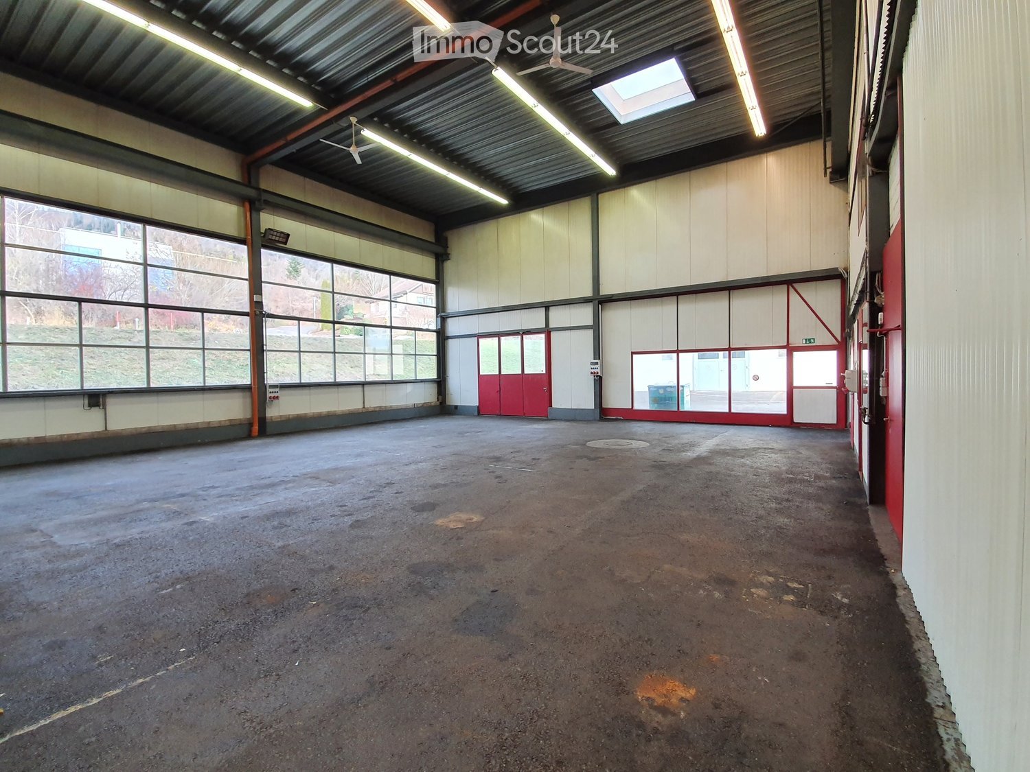 Large, empty industrial-style room with concrete floor, white walls, large windows, and red doors.