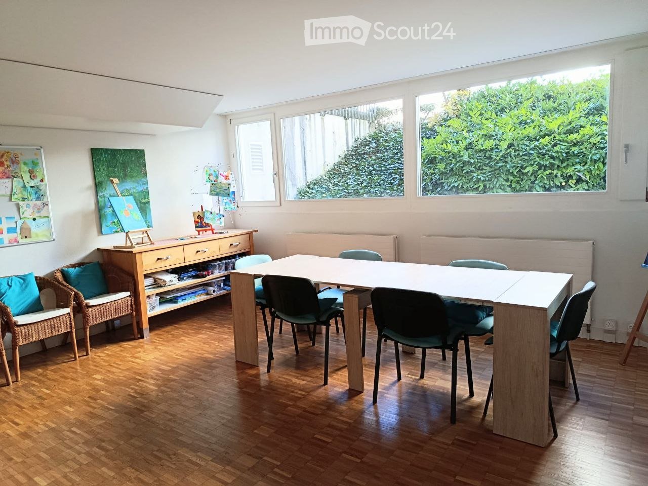 Bright room with wooden flooring, large windows, a long table surrounded by chairs, a wicker sofa with cushions, a wooden cabinet, and several artworks displayed on the walls.