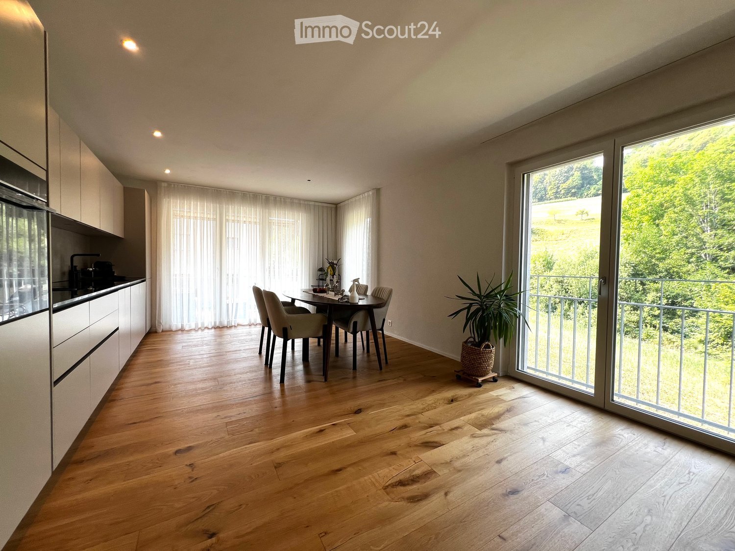 Open-plan kitchen and dining area with light wood flooring, modern kitchen cabinets, a dining table with chairs, and a large window leading to a balcony with a view of greenery.