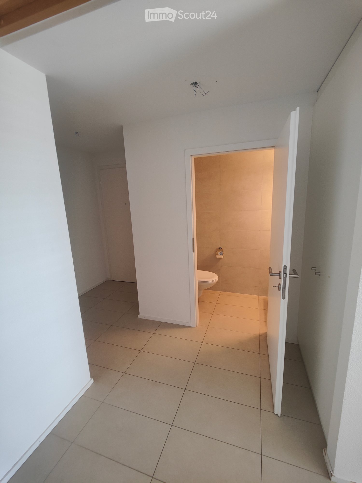 Empty hallway with tiled floor and white walls, leading to a small bathroom with a toilet and tiled walls.