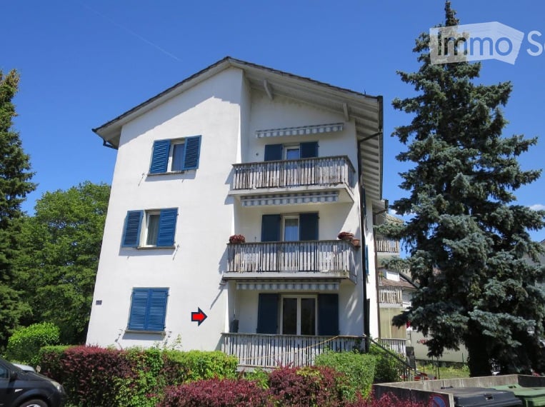 A three-story white building with blue shutters and balconies, surrounded by greenery and trees.