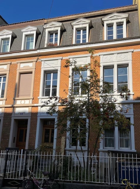 A three-story brick building with white decorative trim around the windows and roofline. The facade features multiple windows with white frames and shutters, and a small tree is planted in front of the building.