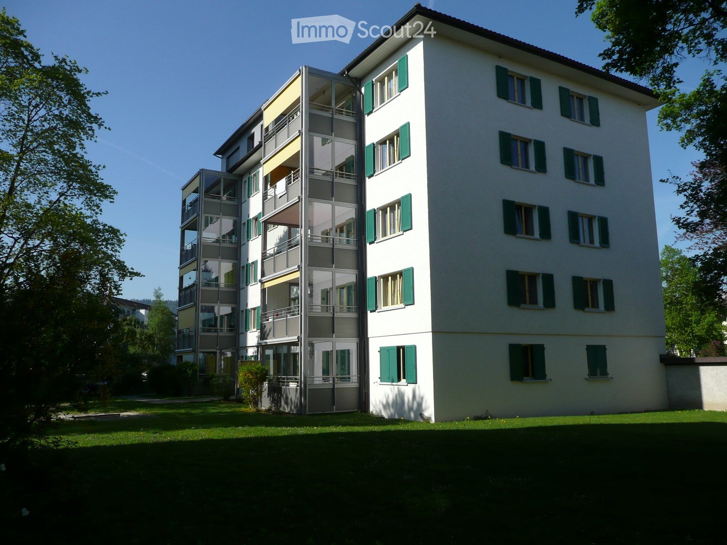 Modern multi-story residential building with white facade, green shutters, and balconies. The building features a mix of glass and concrete elements, surrounded by a well-maintained lawn and trees.