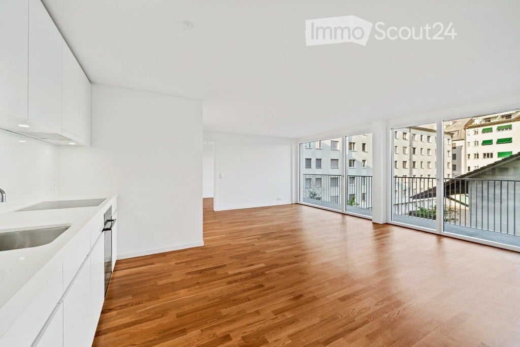 Open-plan unfurnished room with light wooden flooring, white walls, and large sliding glass doors leading to a balcony.