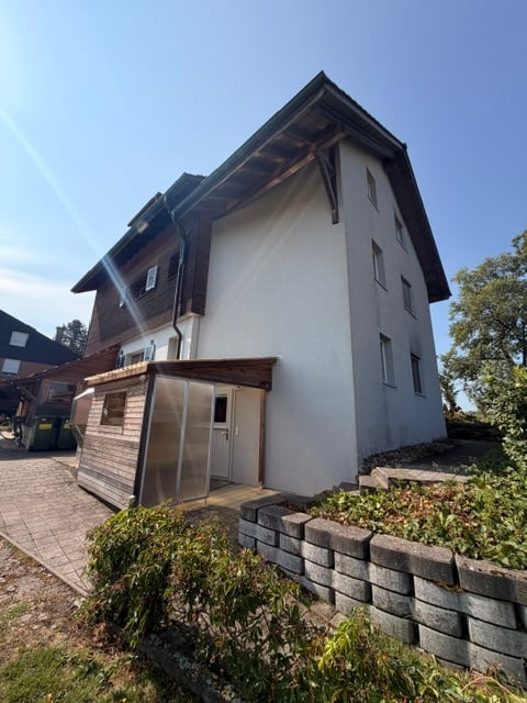 Two-story house with white exterior walls, wooden roof, and a small attached garage. The house features a stone foundation and a landscaped front yard with low shrubs and a paved driveway.