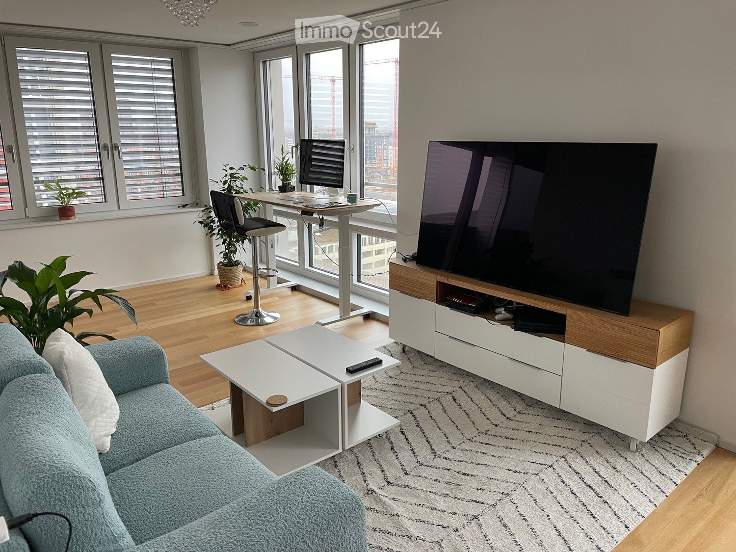 Modern living room with a light blue sofa, white coffee table, TV stand, desk with a stool chair, and plants near the windows.