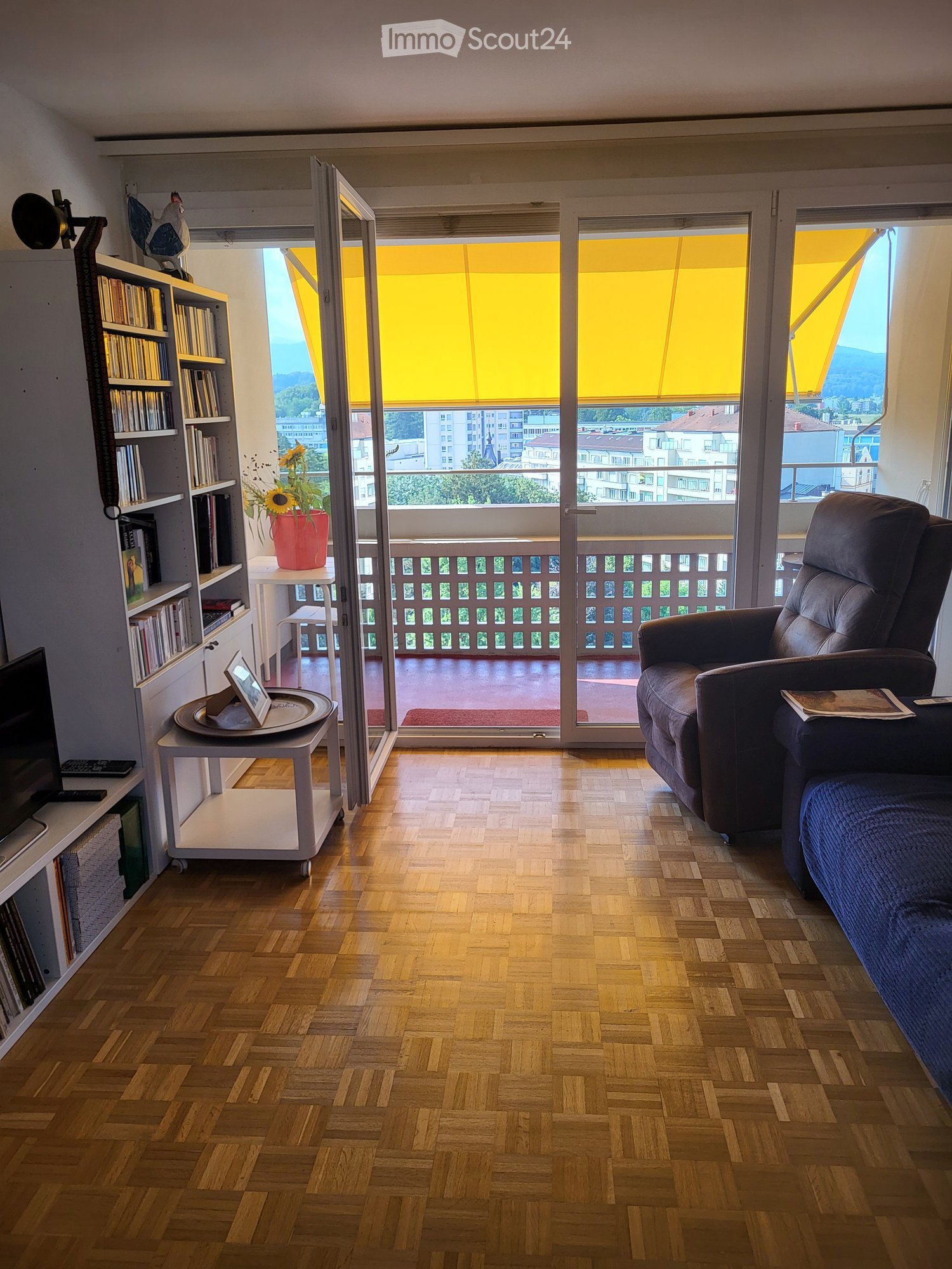 Living room with wooden flooring, large windows, balcony access, sofa, chair, small table, bookshelf, flower pot, and picture frame.