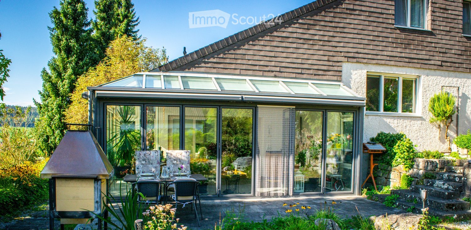 Outdoor patio with a glass-enclosed area, dining table and chairs, surrounded by greenery and a barbecue.