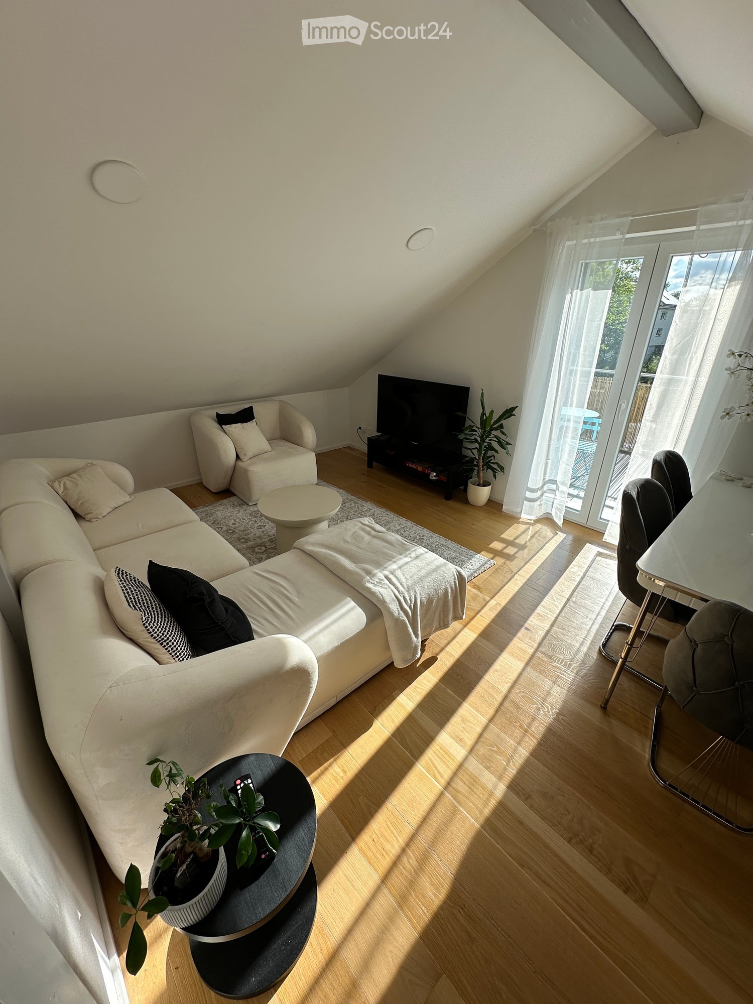 Bright living room with wooden flooring, large windows, and a balcony door. Features a white sectional sofa, armchair, coffee table, TV, and dining area with chairs and table.