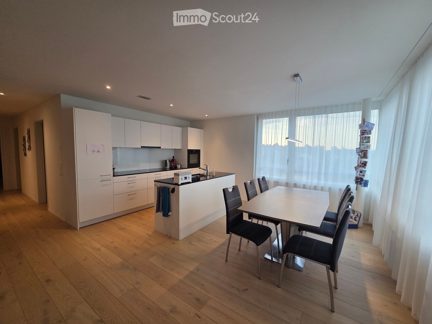 Modern kitchen and dining area with white cabinets, a central island, a dining table with chairs, and large windows with curtains.