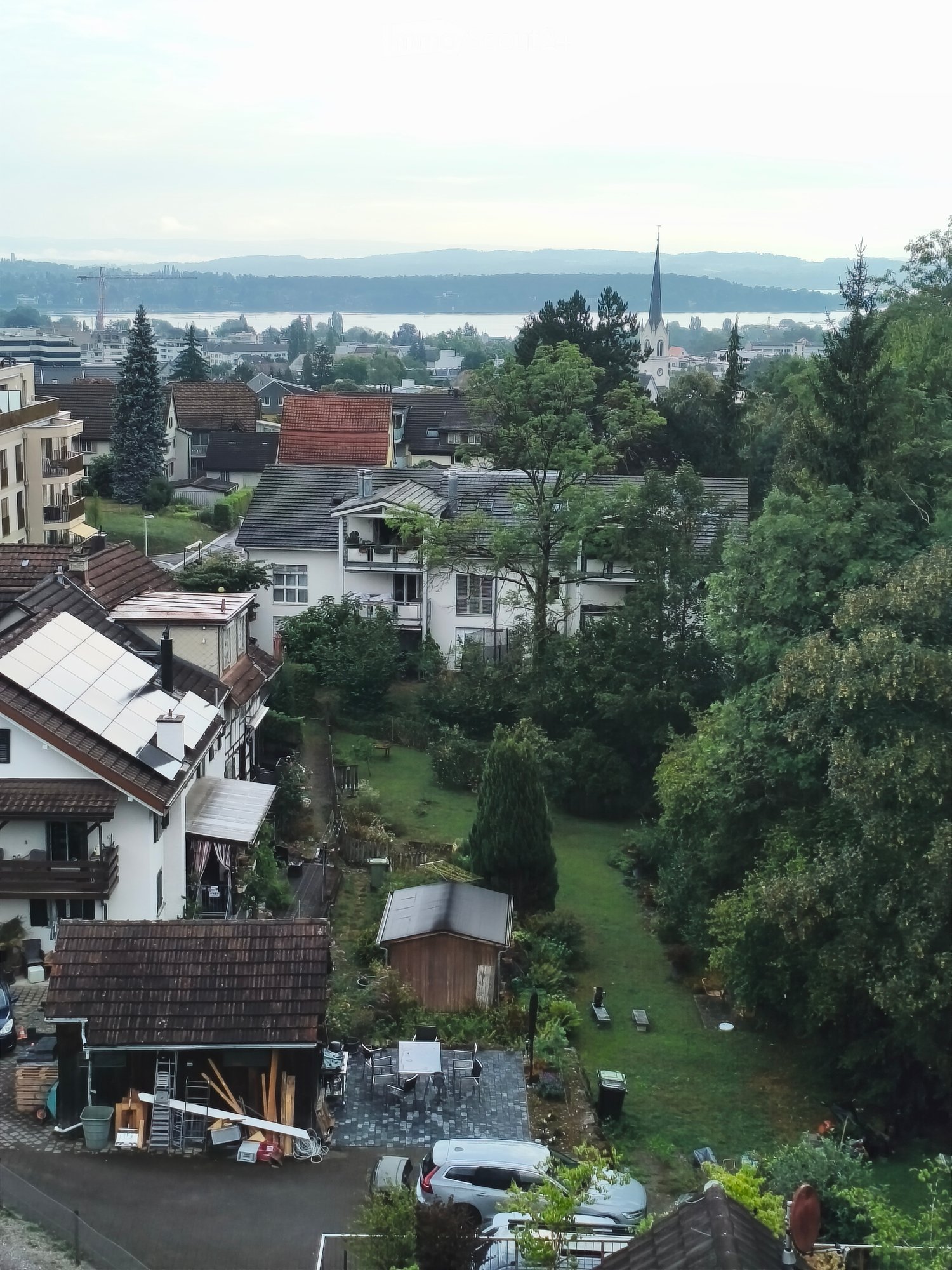 Aerial view of a residential neighborhood with multiple houses, gardens, and parked cars. The area includes a mix of single-family homes with various roof styles and surrounding greenery.