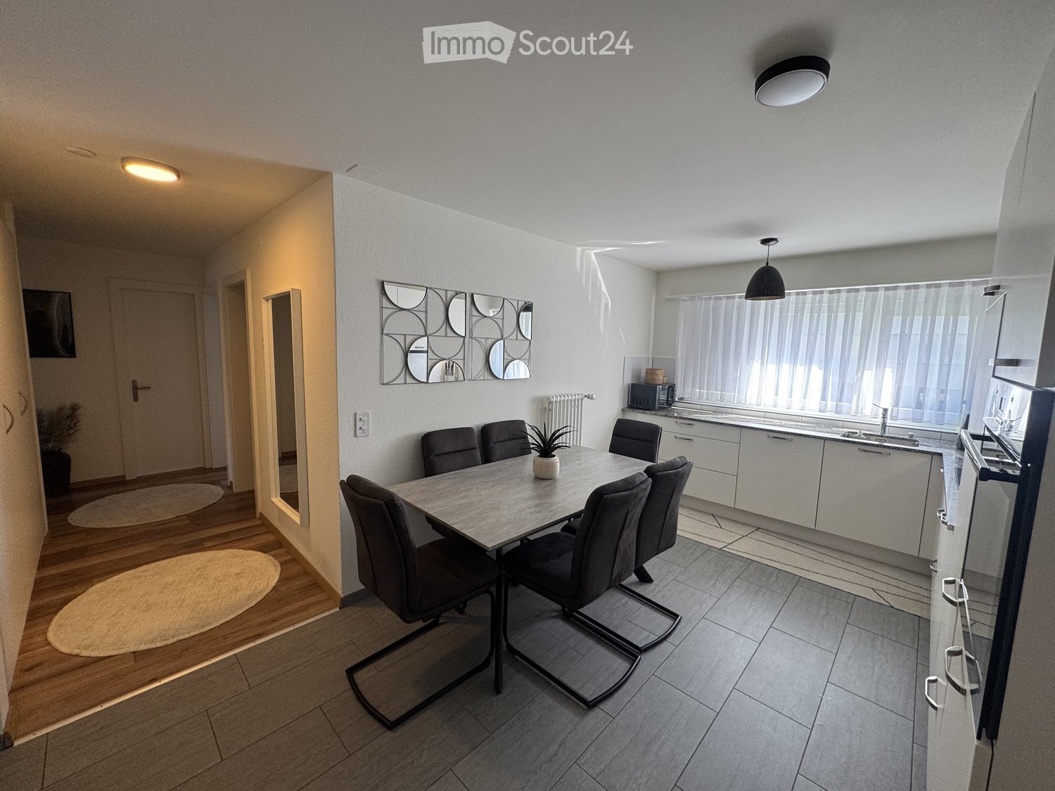 Modern open-plan living and dining area with a dining table, six chairs, a kitchen area with cabinets, sink, microwave, and a decorative mirror on the wall.