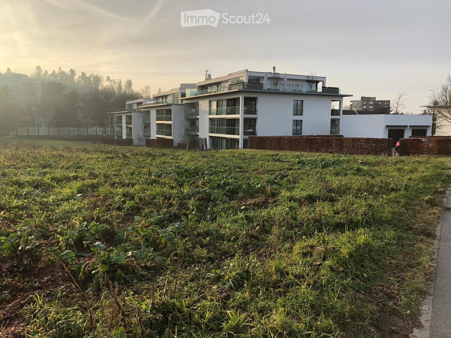 Modern multi-story residential building with large windows and balconies, surrounded by a grassy field and trees in the background.