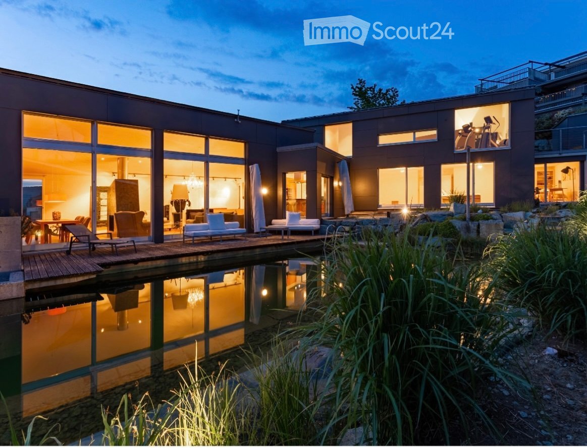Modern two-story house with large glass windows, a reflective water feature, and outdoor seating area.