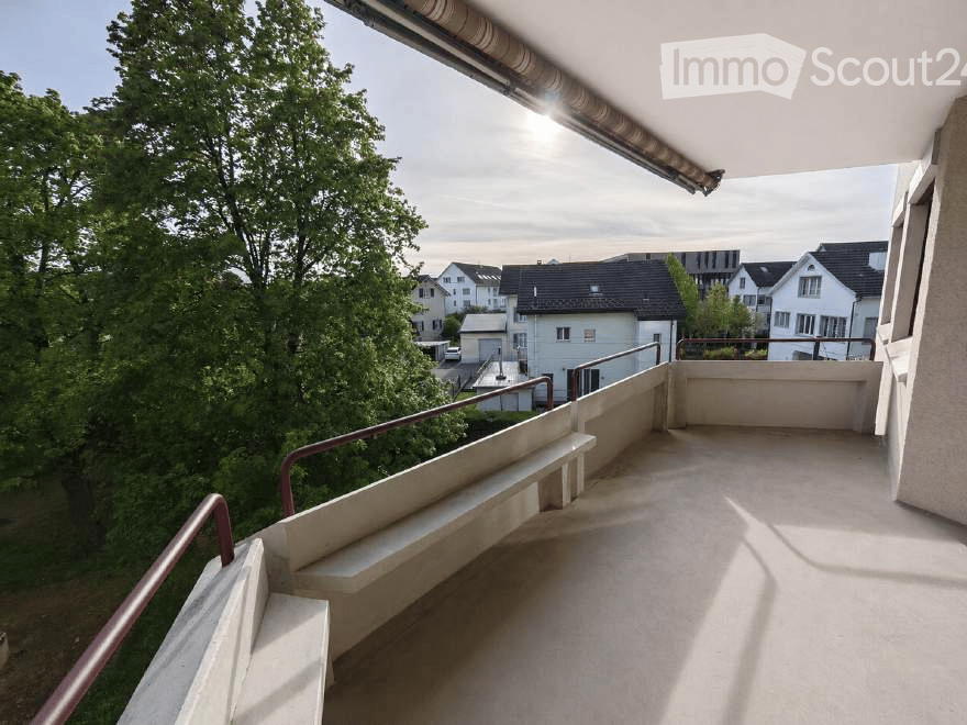 Balcony with concrete floor, metal railing, and steps leading to a lower level, overlooking a residential area with trees and houses.